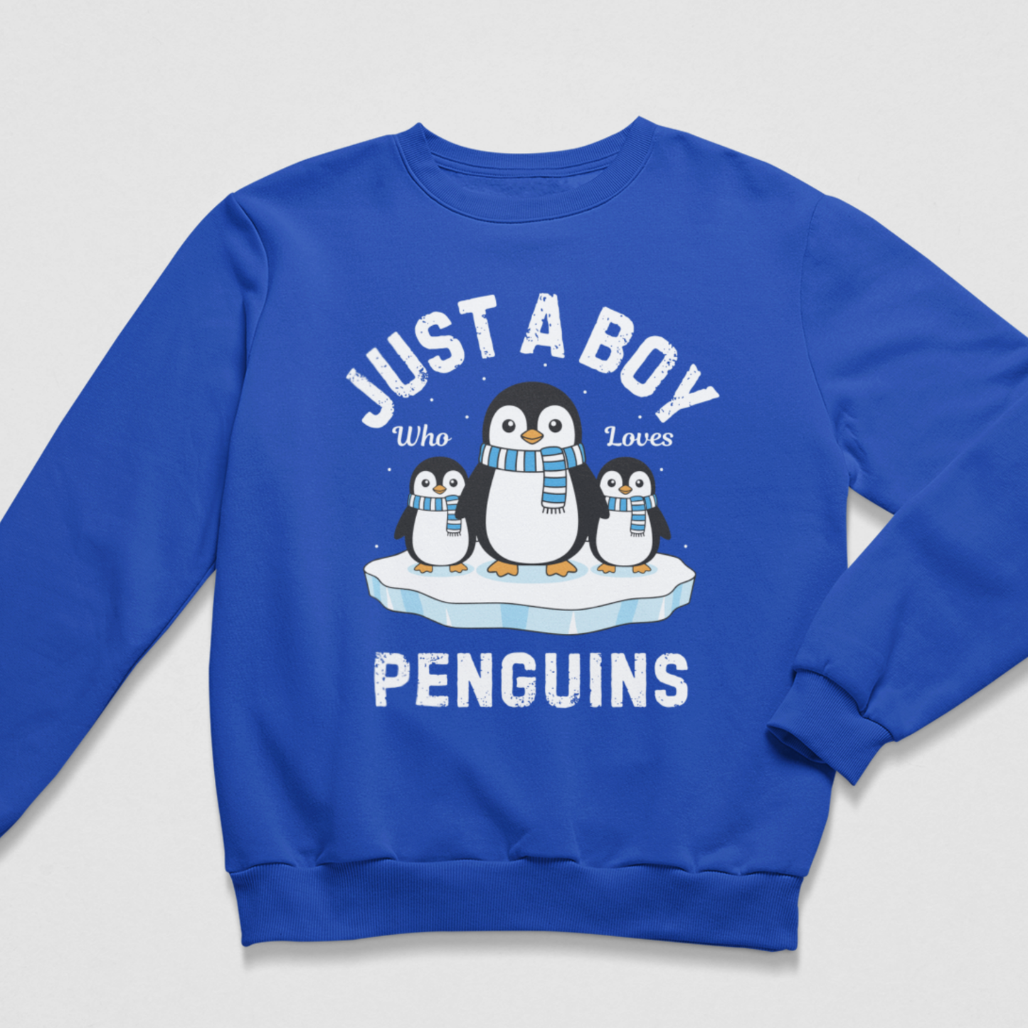 Flat lay of a royal blue crew neck sweatshirt with one large penguin between two smaller penguins, blue-and-white scarves and “Just a Boy Who Loves Penguins” wording.