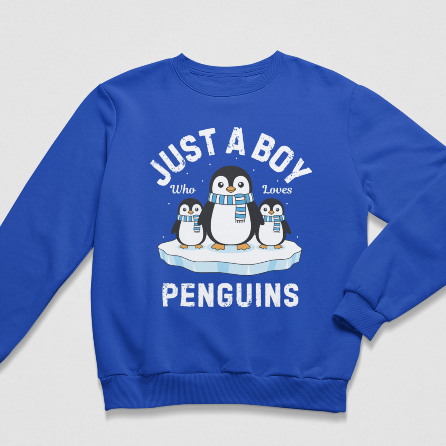 Flat lay of a royal blue crew neck sweatshirt with one large penguin between two smaller penguins, blue-and-white scarves and “Just a Boy Who Loves Penguins” wording.