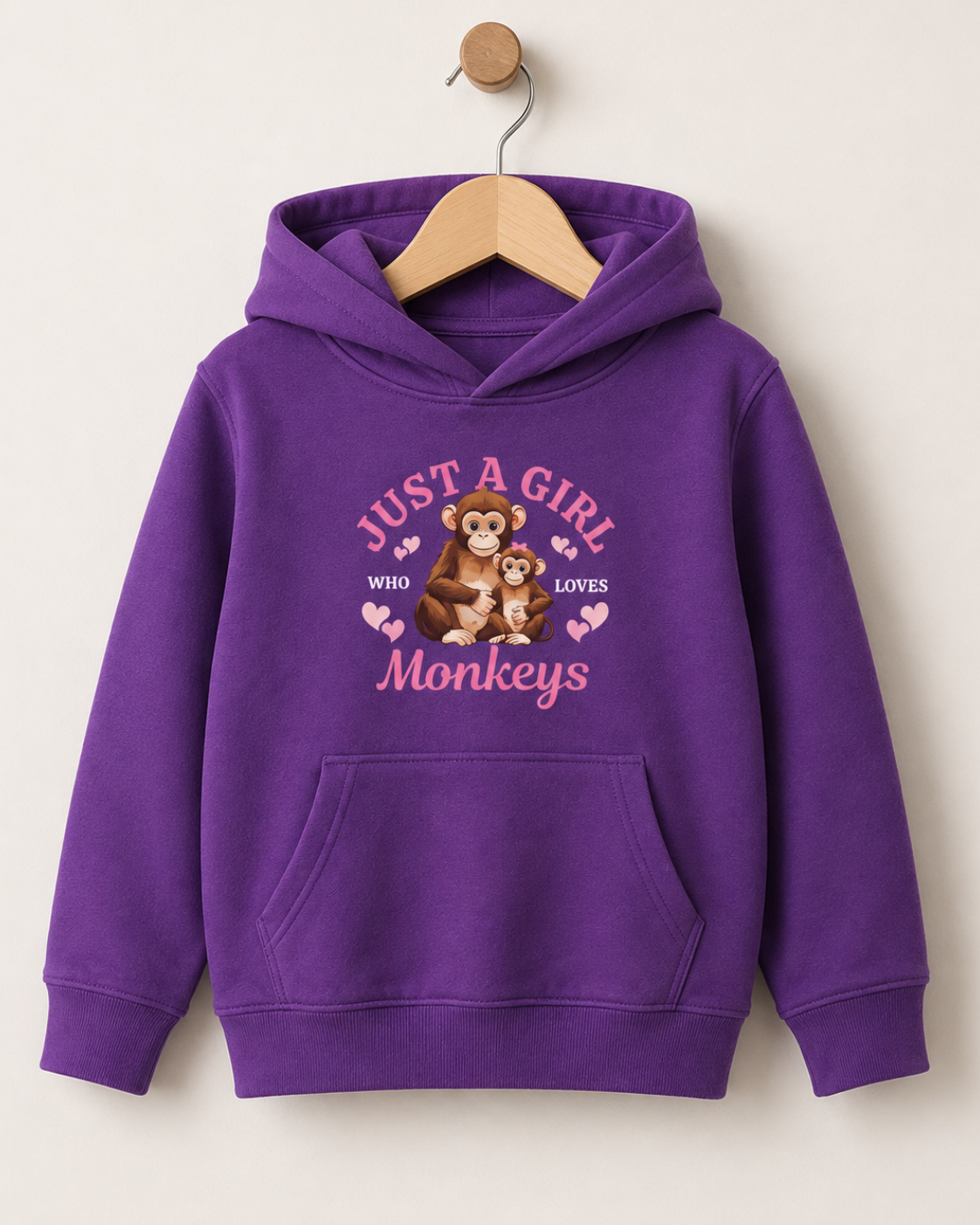 A purple kids pullover hoodie hanging on a wooden hanger against a light beige wall. The hoodie features a front graphic with the text 'JUST A GIRL WHO LOVES Monkeys' in pink and a cartoon monkey illustration.