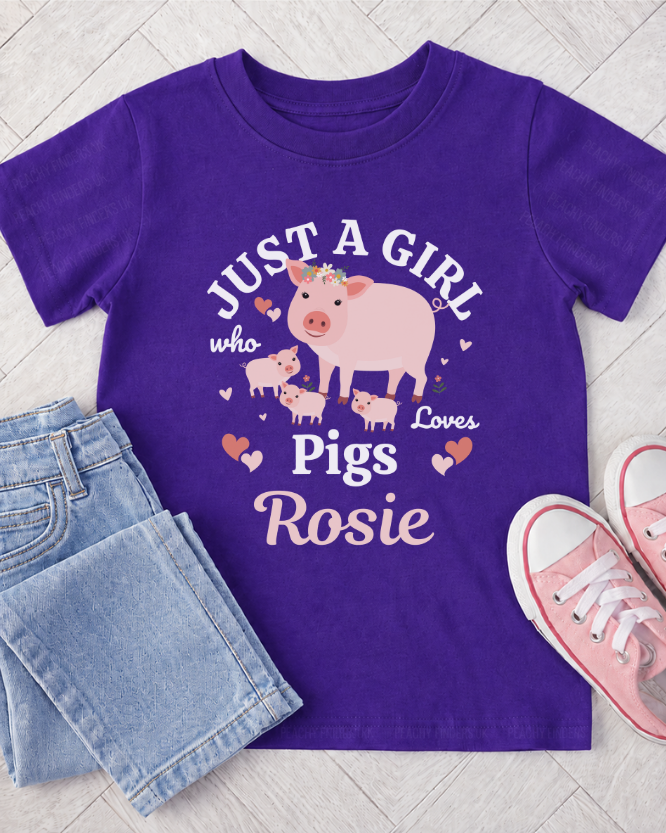 A kids purple short-sleeve t-shirt with a cartoon pig graphic and the text 'JUST A GIRL who Loves Pigs Rosie' in white and pink lettering. The shirt is styled in a flat lay with light blue jeans and pink sneakers on a light grey wood plank background.