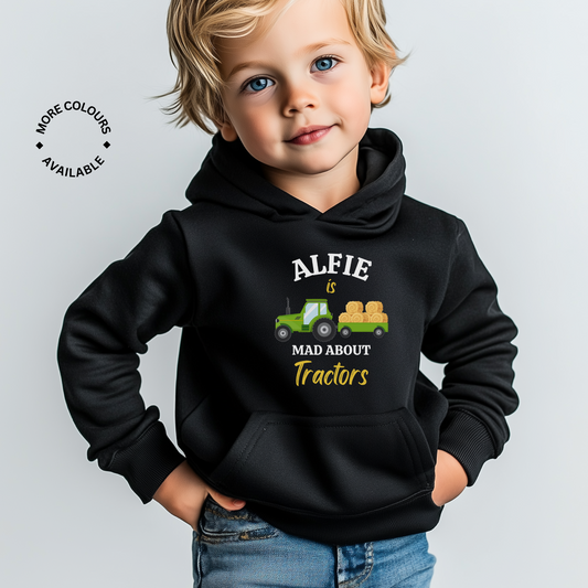 A little boy wearing a black hoodie with a printed tractor and words saying "Alfie is mad about tractors"