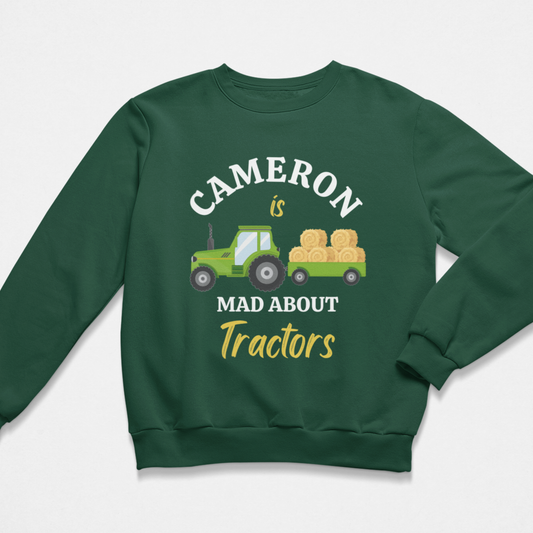 A boys dark green crewneck sweatshirt featuring a graphic print of a green tractor carrying hay bales, with the text 'CAMERON is MAD ABOUT Tractors' in white and yellow lettering.