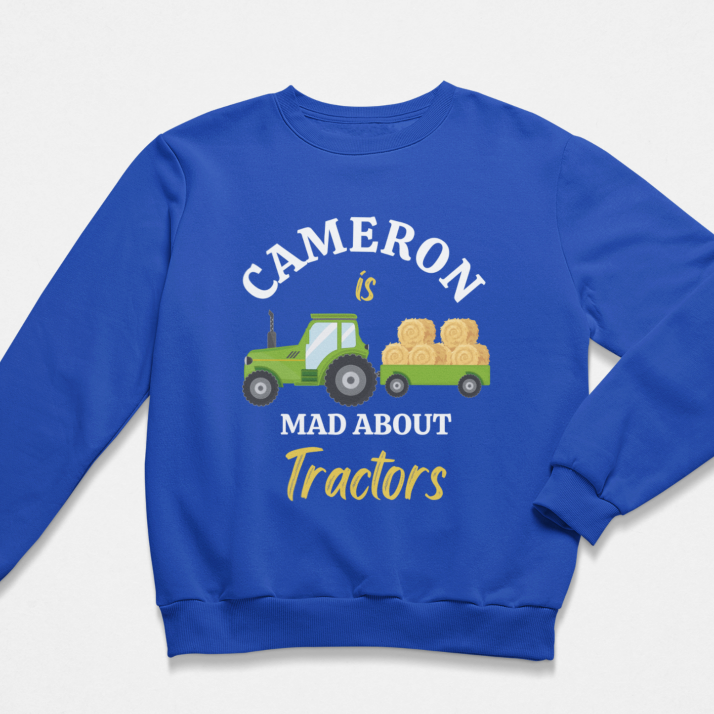 A boys royal blue crewneck sweatshirt featuring a custom graphic print. The design shows a green cartoon tractor pulling a trailer loaded with yellow hay bales, with the text 'CAMERON is MAD ABOUT Tractors' in white and yellow lettering.