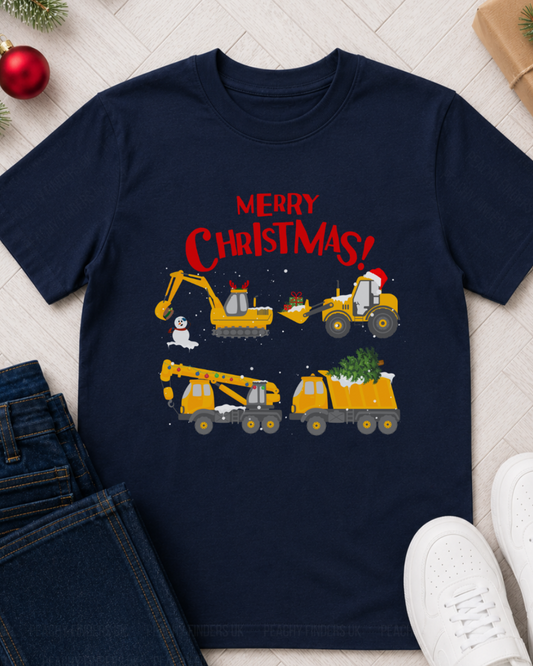 A kids navy blue graphic t-shirt featuring a Christmas construction theme with yellow excavators and trucks, displayed in a flat-lay arrangement with jeans, white sneakers, and holiday decorations on a light wood background.