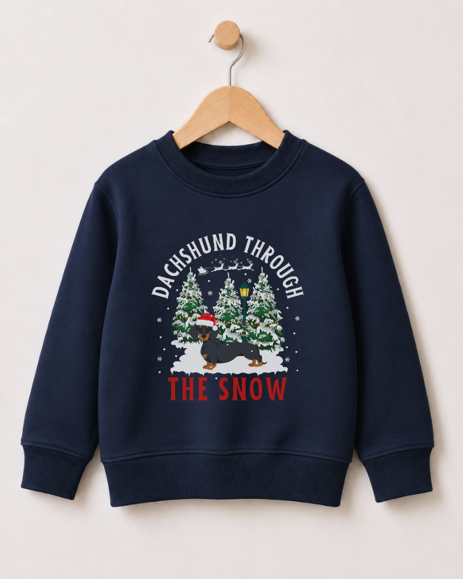 A kids navy blue crewneck sweatshirt hanging on a wooden hanger against a light beige wall. The sweatshirt features a festive graphic with the text 'DACHSHUND THROUGH THE SNOW' in white and red, surrounding an illustration of a dachshund wearing a Santa hat under snow-covered Christmas trees.