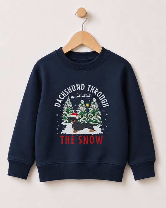 A kids navy blue crewneck sweatshirt hanging on a wooden hanger against a light beige wall. The sweatshirt features a festive graphic with the text 'DACHSHUND THROUGH THE SNOW' in white and red, surrounding an illustration of a dachshund wearing a Santa hat under snow-covered Christmas trees.