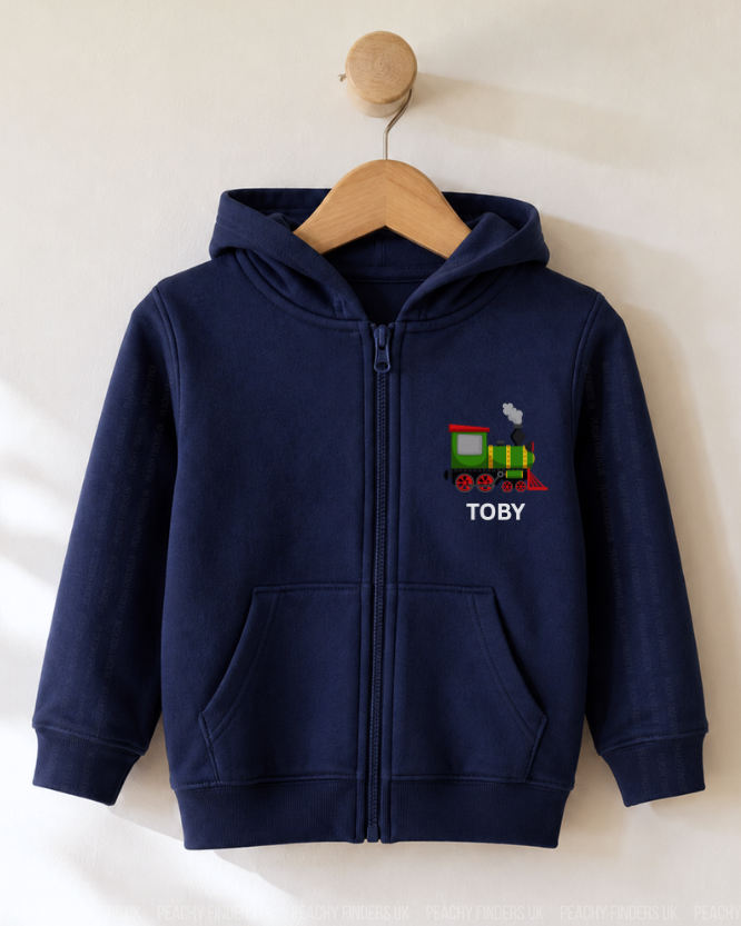A navy blue zip-up hoodie for a child, hanging on a wooden hanger against a light beige wall. The hoodie features a colourful printed train graphic and the name 'TOBY' in white text on the left chest.