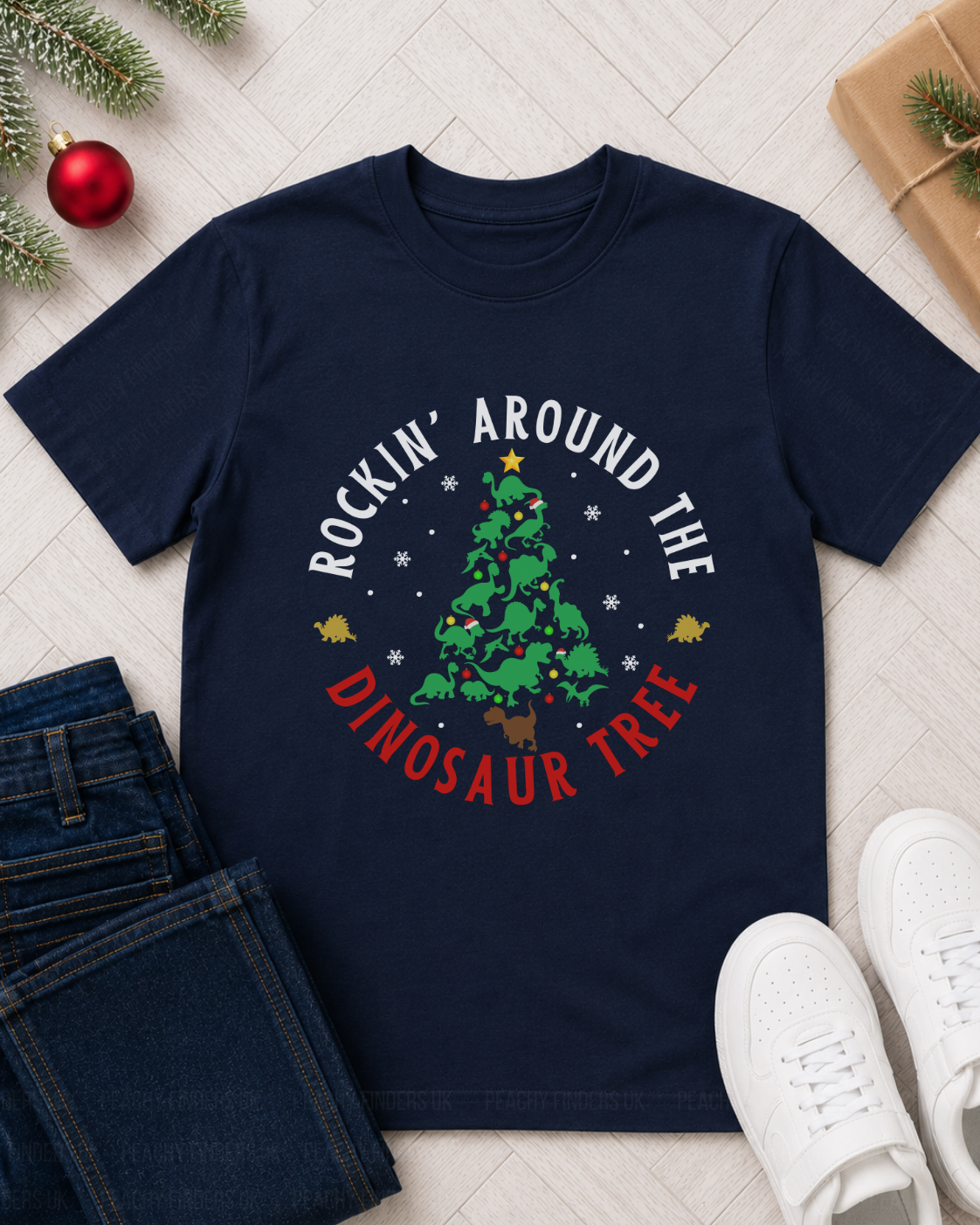A children's navy blue graphic t-shirt featuring a Christmas tree design with the text 'ROCKIN' AROUND THE DINOSAUR TREE' in white and red. The shirt is styled in a flat-lay arrangement on a light wood floor alongside folded blue jeans, white sneakers, a red ornament, and a wrapped gift box.