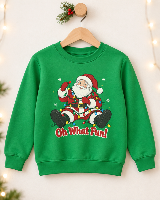 Kids kelly green “Oh What Fun!” Santa sweatshirt on a wooden hanger, with a pine sprig and softly blurred fairy lights.