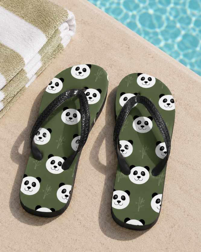 A pair of green flip-flop sandals with a black strap and a repeating panda face and bamboo pattern, placed on a beige poolside deck with a folded towel and blue water in the background.