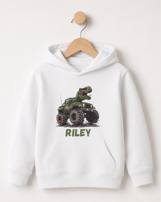 Kids hoodie in white with T-Rex riding camouflage monster truck and green personalised name Riley on the front styled on a wooden hanger.