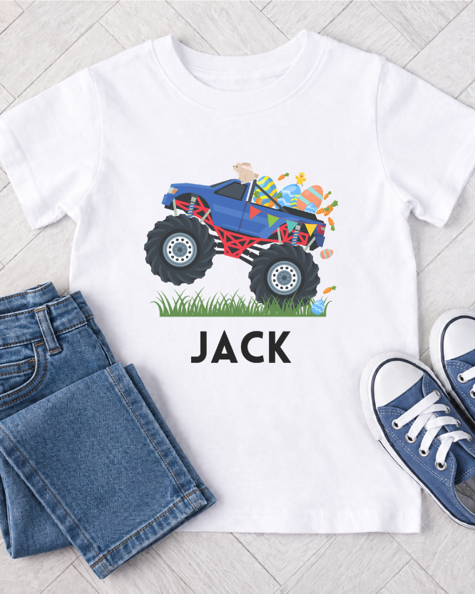 Kids white t-shirt with a colourful monster truck graphic and custom name 'JACK' text, paired with blue jeans and shoes on a light wooden floor.