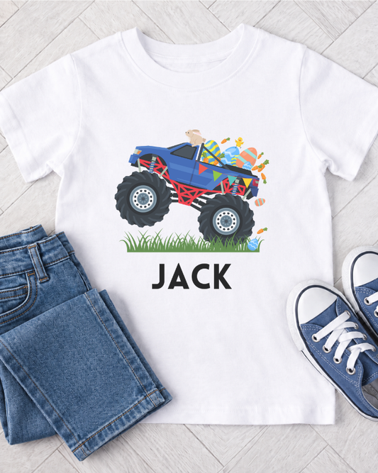Kids white t-shirt with a colourful monster truck graphic and custom name 'JACK' text, paired with blue jeans and shoes on a light wooden floor.