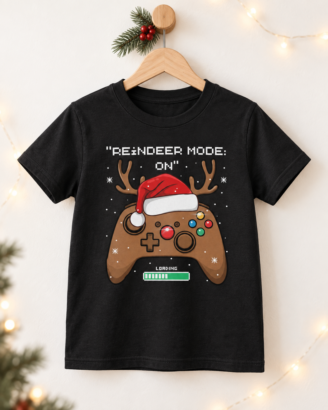 Black crewneck kids’ Christmas T-shirt with a Santa-hat gaming controller, displayed on a wooden hanger with holly and warm lights.