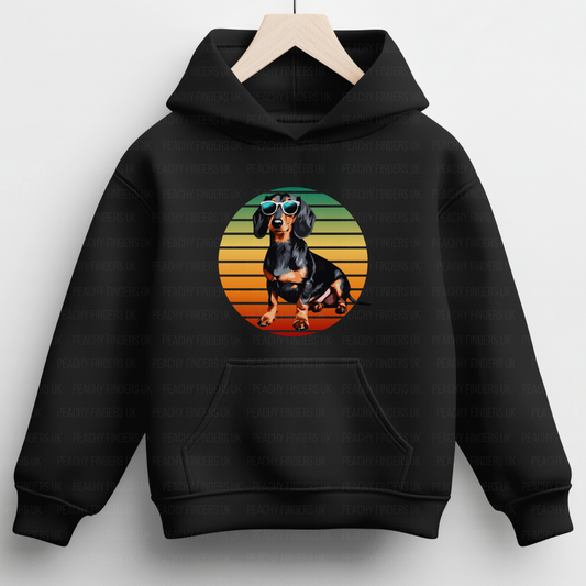 A kids black hooded sweatshirt hangs on a wooden hanger. The hoodie features a circular graphic of a black and tan Dachshund dog wearing mirrored sunglasses, set against horizontal stripes sunset of orange, yellow, and green.