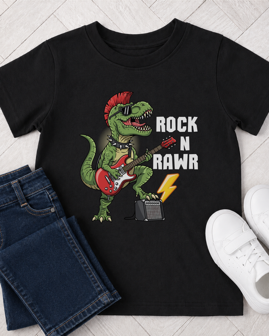 A kids black short-sleeve graphic T-shirt laid flat on a light wood floor, styled with folded blue jeans on the left and white sneakers on the right. The shirt features a green dinosaur playing a red electric guitar with the text 'ROCK N RAWR' in white.