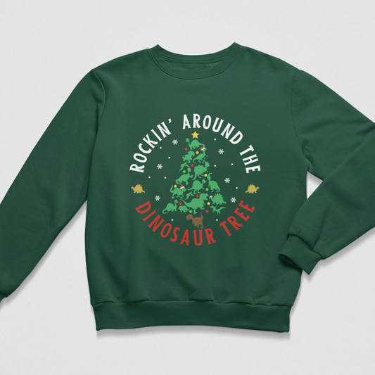 A kids dark green crewneck sweatshirt featuring a circular graphic on the chest with a green dinosaur Christmas tree, white text reading 'ROCKIN' AROUND THE', and red text reading 'DINOSAUR TREE' on a light grey background.