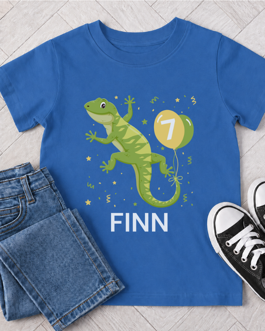 A royal blue kids t-shirt with a green gecko and a yellow balloon graphic, personalised with the name 'FINN' in white. The shirt is styled in a flat lay with blue jeans and black sneakers on a light grey wood floor.
