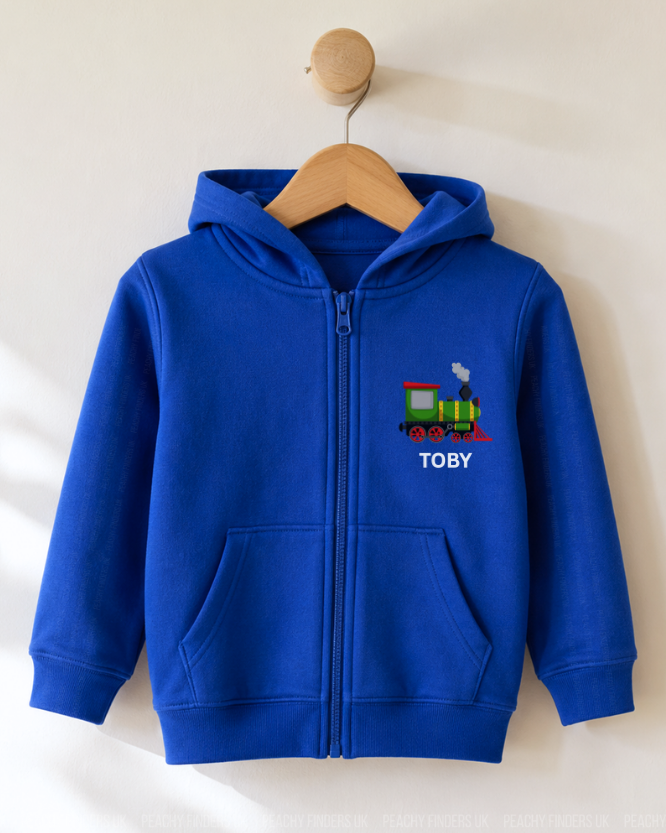 A royal blue zip-up hoodie for toddlers, hanging on a wooden hanger against a warm off-white wall. The hoodie features a colourful printed train graphic and the name 'TOBY' in white text on the left chest.
