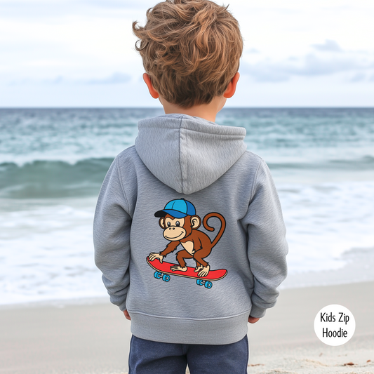Young boy wearing grey kids zip hoodie with a cartoon monkey riding a red skateboard printed on the back, standing on the beach