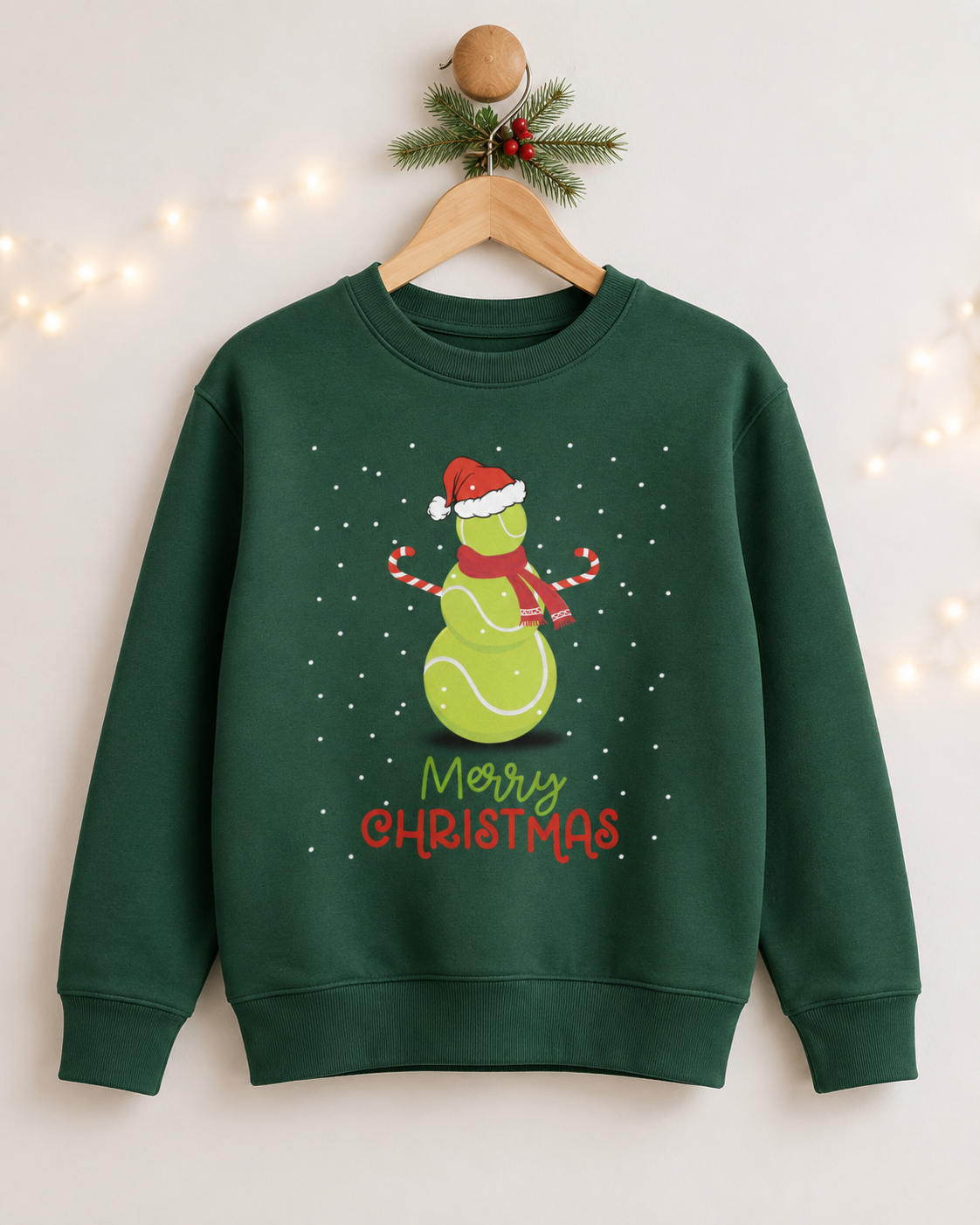 A kids dark green crewneck sweatshirt hanging on a wooden hanger decorated with a pine branch and fairy lights. The sweatshirt features a graphic of a green tennis ball snowman wearing a red Santa hat and scarf, with 'Merry Christmas' written below in green and red.