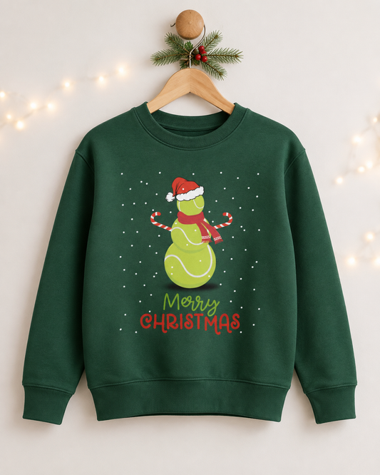 A kids dark green crewneck sweatshirt hanging on a wooden hanger decorated with a pine branch and fairy lights. The sweatshirt features a graphic of a green tennis ball snowman wearing a red Santa hat and scarf, with 'Merry Christmas' written below in green and red.