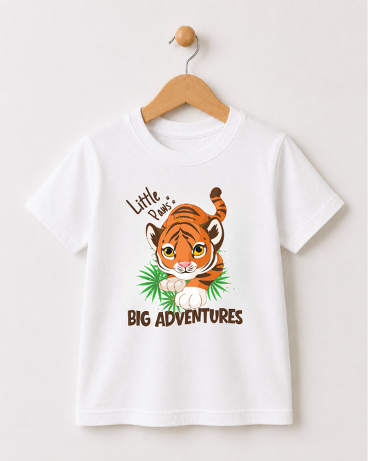 A white short-sleeve kids t-shirt hanging on a wooden hanger against a light beige wall. The shirt features a cartoon graphic of a cute orange and brown striped tiger walking on green leaves, with the text 'Little Paw's' above and 'BIG ADVENTURES' below.