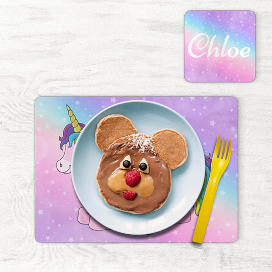 Kids personalised unicorn placemat and coaster set with pastel star design and the name “Chloe” in white script. A plate with bear-shaped pancakes and a yellow fork are on the placemat.