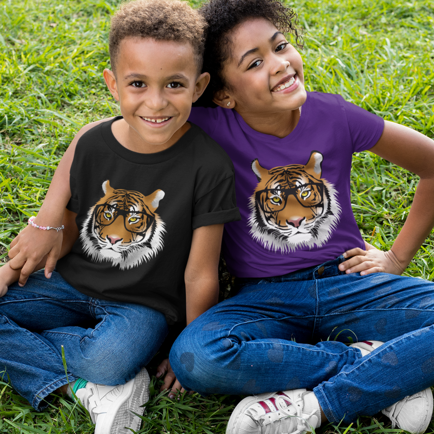 A boy and a girl sitting on grass wearing kids t-shirts featuring a tiger wearing glasses - one in black and one in purple - showing a fun, clever animal design.