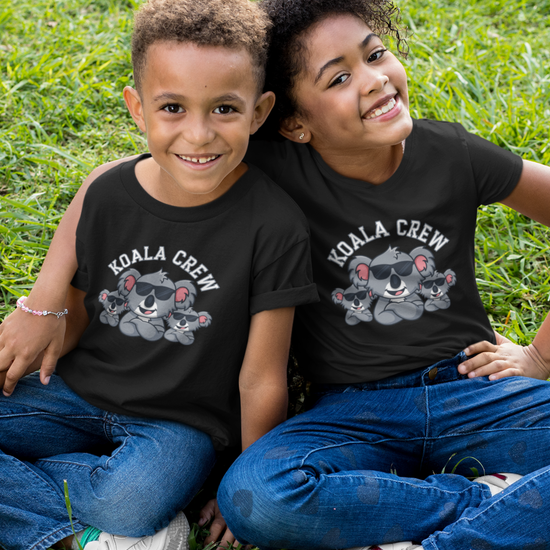 Two smiling children sitting on grass wearing matching black Koala Crew T-shirts with cartoon koala family graphic and text – fun cotton kids tee for ages 3–13.