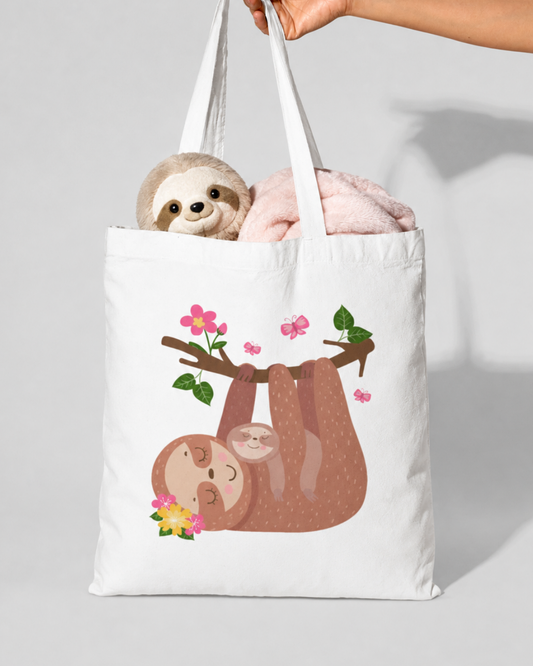 Kids white cotton tote bag featuring a mum and baby sloth hanging from a floral branch, filled with a small plush sloth toy and a soft light pink blanket.