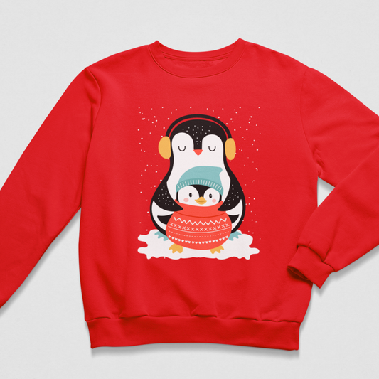 A kids red crewneck sweatshirt featuring a cute cartoon penguin graphic on the front. The penguin wears yellow ear muffs, a light blue knit hat, and holds a red patterned sweater while standing on white snow against a snowy background.
