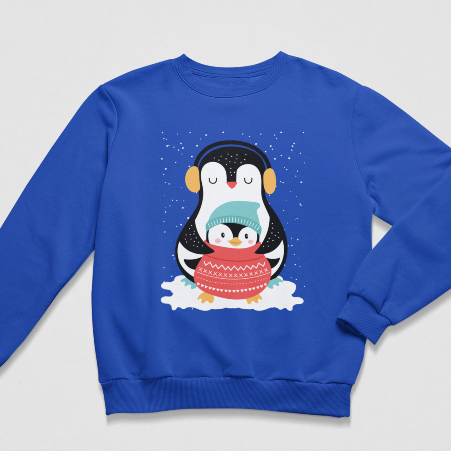 A kids royal blue crewneck sweatshirt featuring a cute cartoon penguin graphic on the chest. The penguin illustration shows a larger penguin wearing black earmuffs and a smaller one hugging it from the front in a red knit sweater, set against a snowy white background with light blue snowflakes.