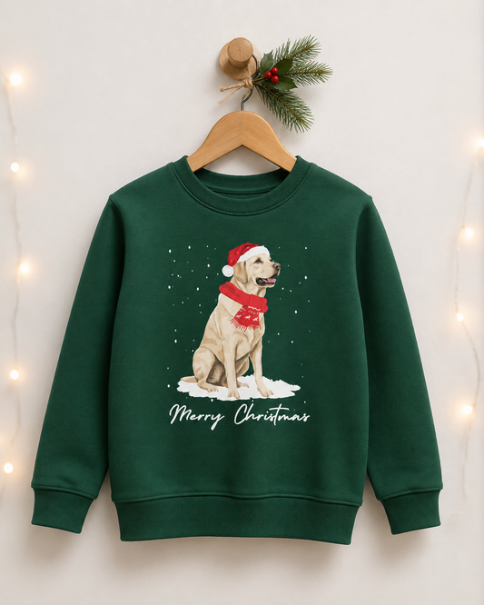 A kids dark green crewneck sweatshirt hanging on a wooden hanger against a light wall, decorated with warm fairy lights and a pine sprig. The sweatshirt features a graphic of a yellow Labrador retriever wearing a red Santa hat and scarf, sitting in snow with 'Merry Christmas' written below.