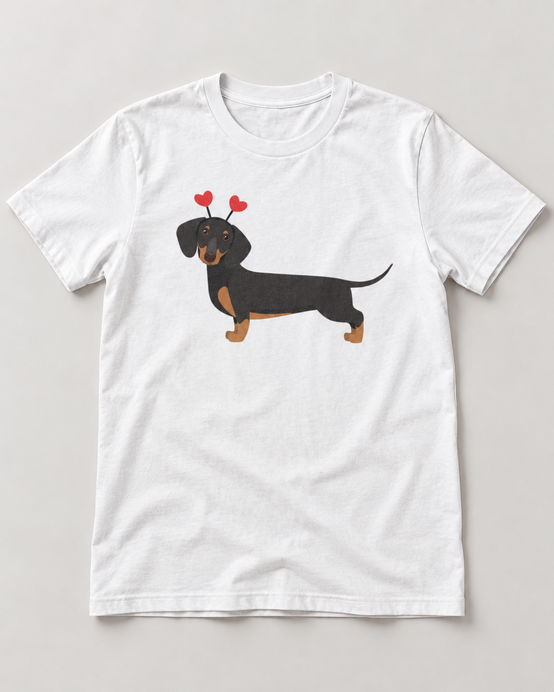 A white short-sleeve crewneck t-shirt featuring a cartoon illustration of a black and tan dachshund wearing red heart-shaped headband on the chest, displayed flat against a light grey background.