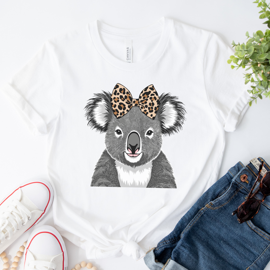 Women’s white cotton t-shirt featuring a cute grey koala with a leopard print bow, styled flat with denim shorts, white sneakers, and sunglasses.
