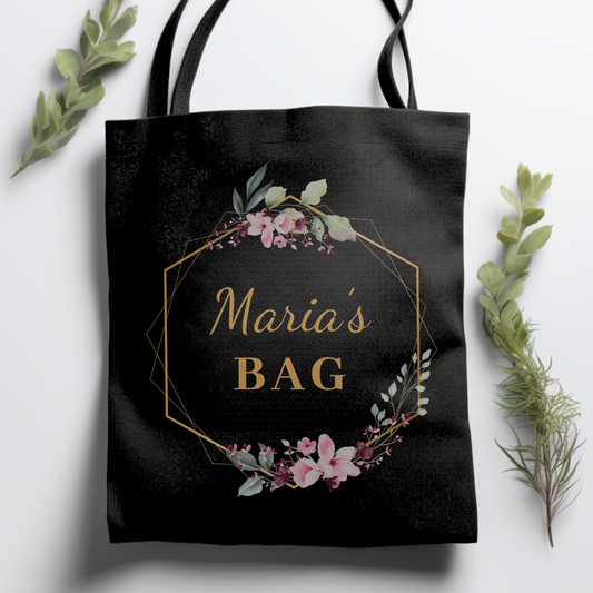 A black cotton tote bag with gold floral hexagon frame and writing on the front.