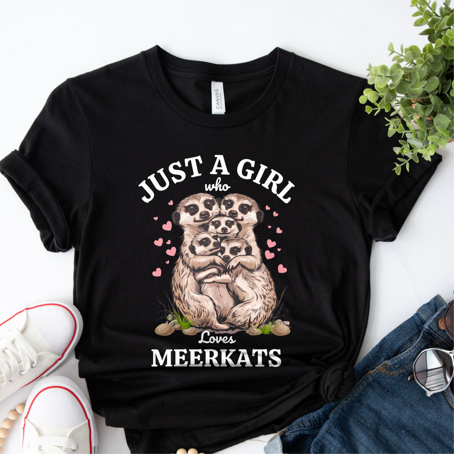 Flat lay of a black women’s t-shirt with “Just a Girl Who Loves Meerkats” design featuring a group of cuddling meerkats and pink hearts, styled with jeans, sunglasses and white trainers.
