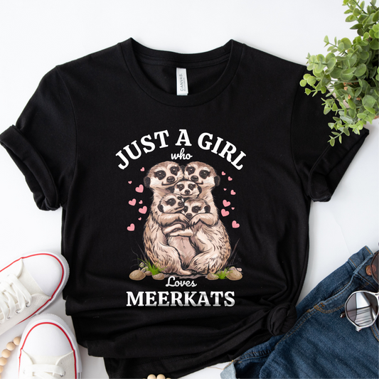 Flat lay of a black women’s t-shirt with “Just a Girl Who Loves Meerkats” design featuring a group of cuddling meerkats and pink hearts, styled with jeans, sunglasses and white trainers.