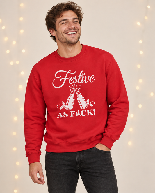 A smiling man laughing while wearing a bright red adult Christmas crewneck sweatshirt featuring a white graphic of clinking beer bottles and the text "FESTIVE AS FUCK!", posed against a softly lit background with string lights.