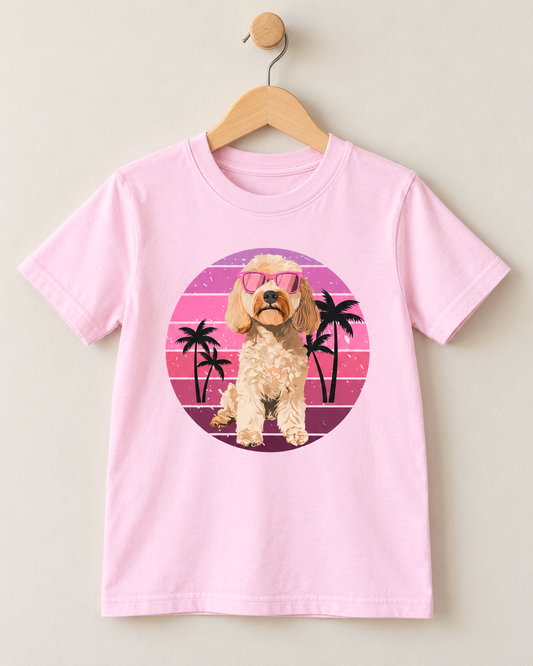 A light pink short-sleeve t-shirt hanging on a wooden hanger against a neutral beige wall. The shirt features a circular graphic print of a cockapoo dog wearing pink sunglasses, set against a retro sunset background with pink and purple stripes and black palm trees.