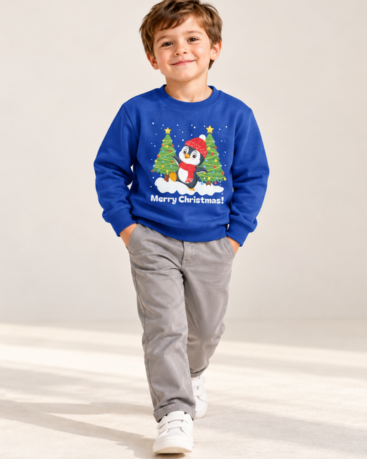 Smiling boy wearing a royal blue penguin Christmas jumper with grey trousers and white trainers, hands in his pockets in a bright studio.
