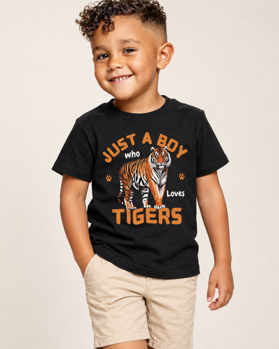 A young boy wearing a black graphic t-shirt with an orange and white tiger design and the text 'JUST A BOY who Loves TIGERS', paired with khaki shorts, against a plain light beige background.
