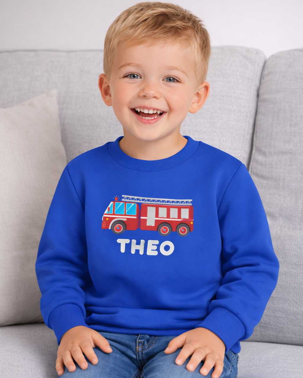 Kids personalised fire truck sweatshirt with name Theo in royal blue, lifestyle photo on sofa