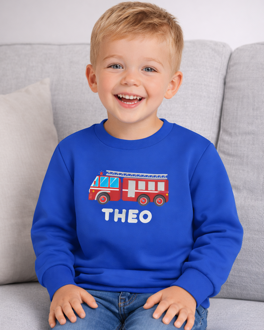 Kids personalised fire truck sweatshirt with name Theo in royal blue, lifestyle photo on sofa