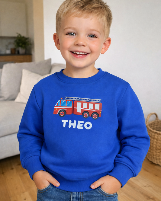Kids personalised fire truck sweatshirt in royal blue with name Theo, worn with denim jeans in living room setting