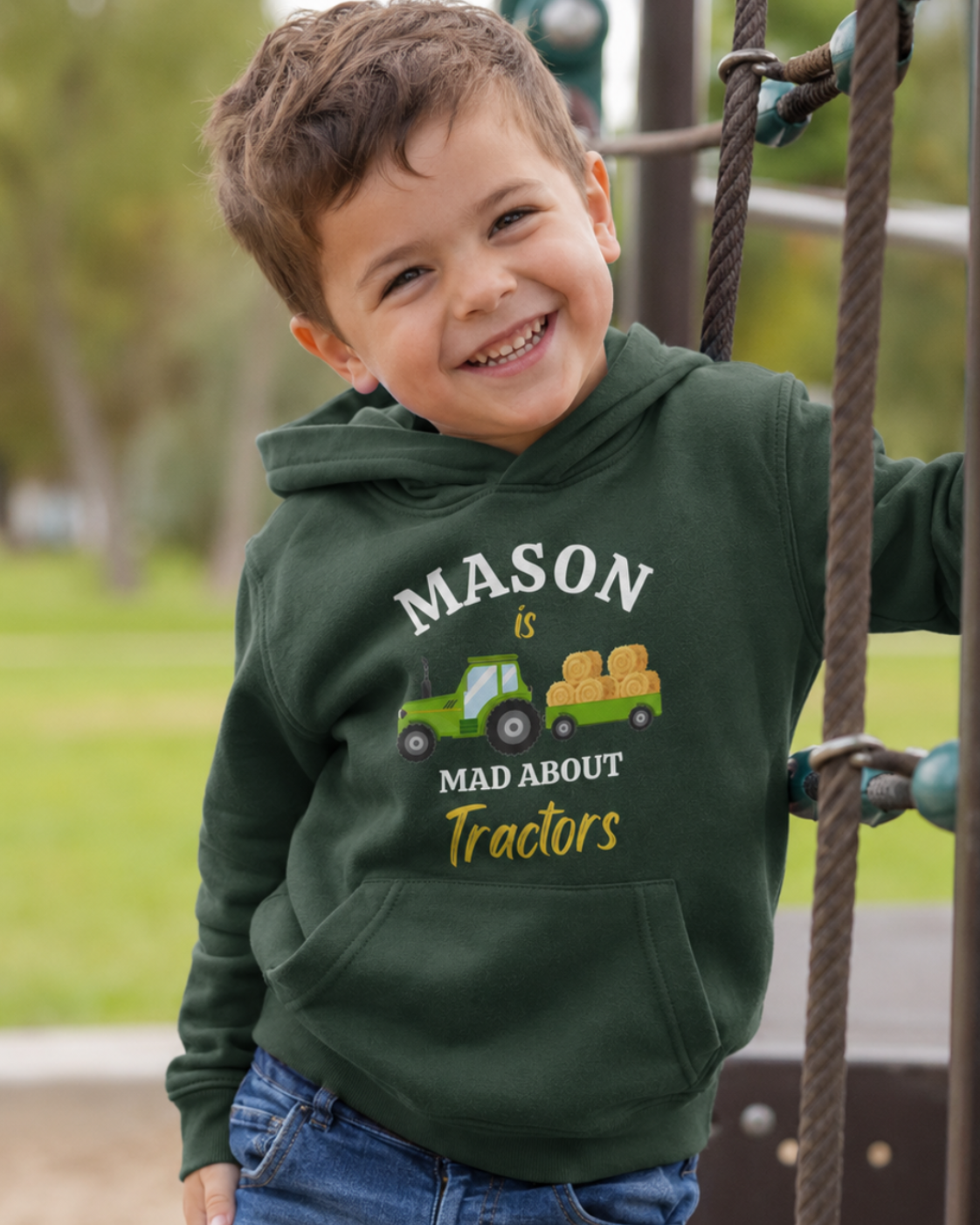 A young boy smiling while climbing playground ropes, wearing a dark green custom hoodie with a graphic that reads 'MASON is MAD ABOUT Tractors' with a green tractor and yellow hay bale.