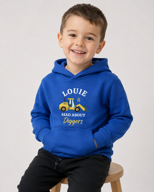 A smiling young boy wearing a royal blue custom hoodie with a yellow construction truck graphic and the text 'LOUIE IS MAD ABOUT Diggers', sitting on a wooden stool against a studio background.