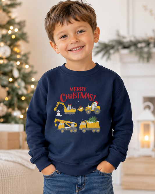 A young boy smiling in a cozy holiday setting, wearing a navy blue crewneck sweatshirt with a 'Merry Christmas' graphic featuring yellow construction vehicles and Santa hat, paired with blue jeans.