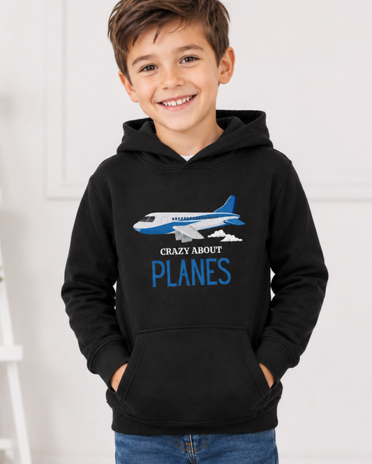 A smiling young boy wearing a black pullover hoodie with a graphic of a blue and white airplane and the text 'CRAZY ABOUT PLANES' in blue, paired with blue jeans against a bright indoor background.
